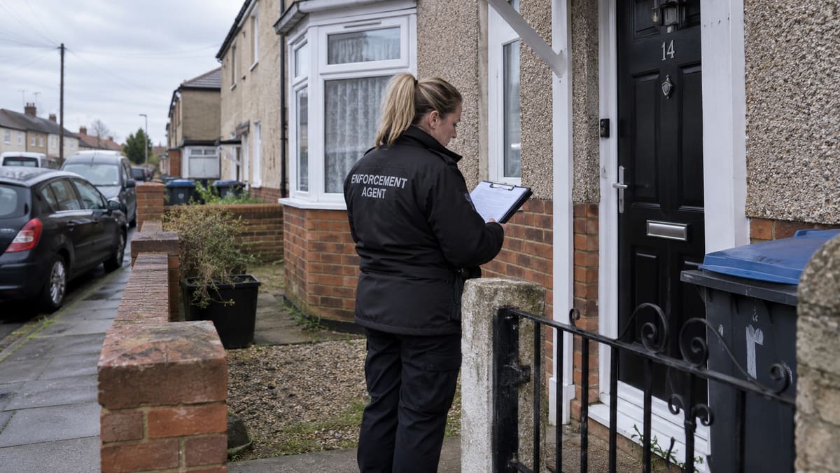 An enforcement agent in a black jacket marked Enforcement Agent reads paperwork on a clipboard outside the front door of a house.